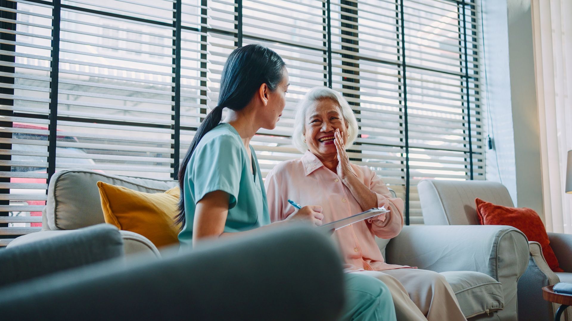 Caregiver and senior woman talking on a sofa. The caregiver is holding a clipboard.