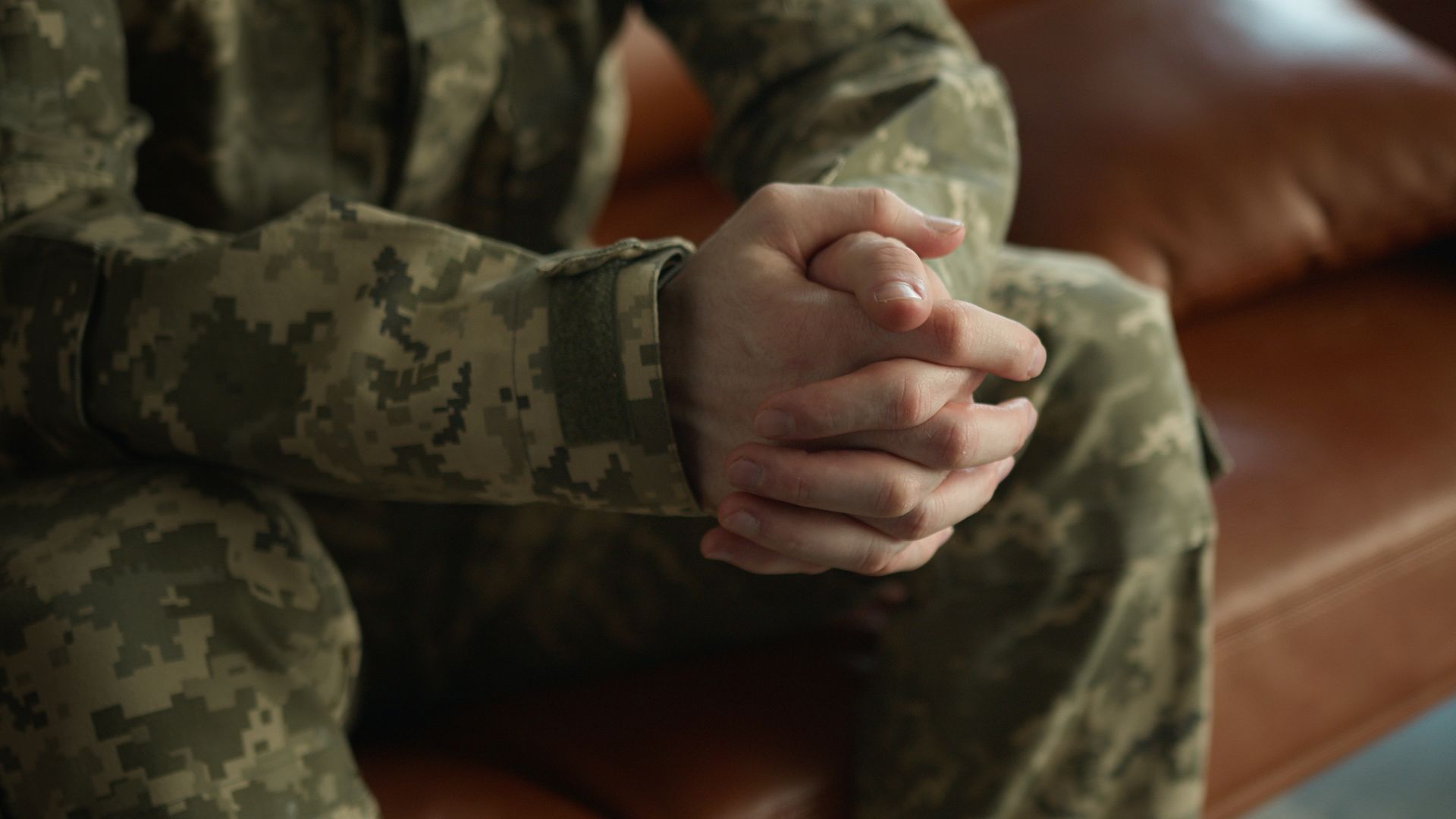 A person in camouflage uniform sits with clasped hands on their lap.