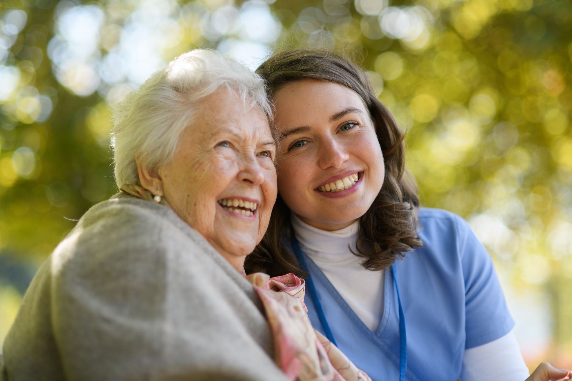 An elderly woman and a younger woman smile outdoors. The younger woman has her arm around the elderly woman.