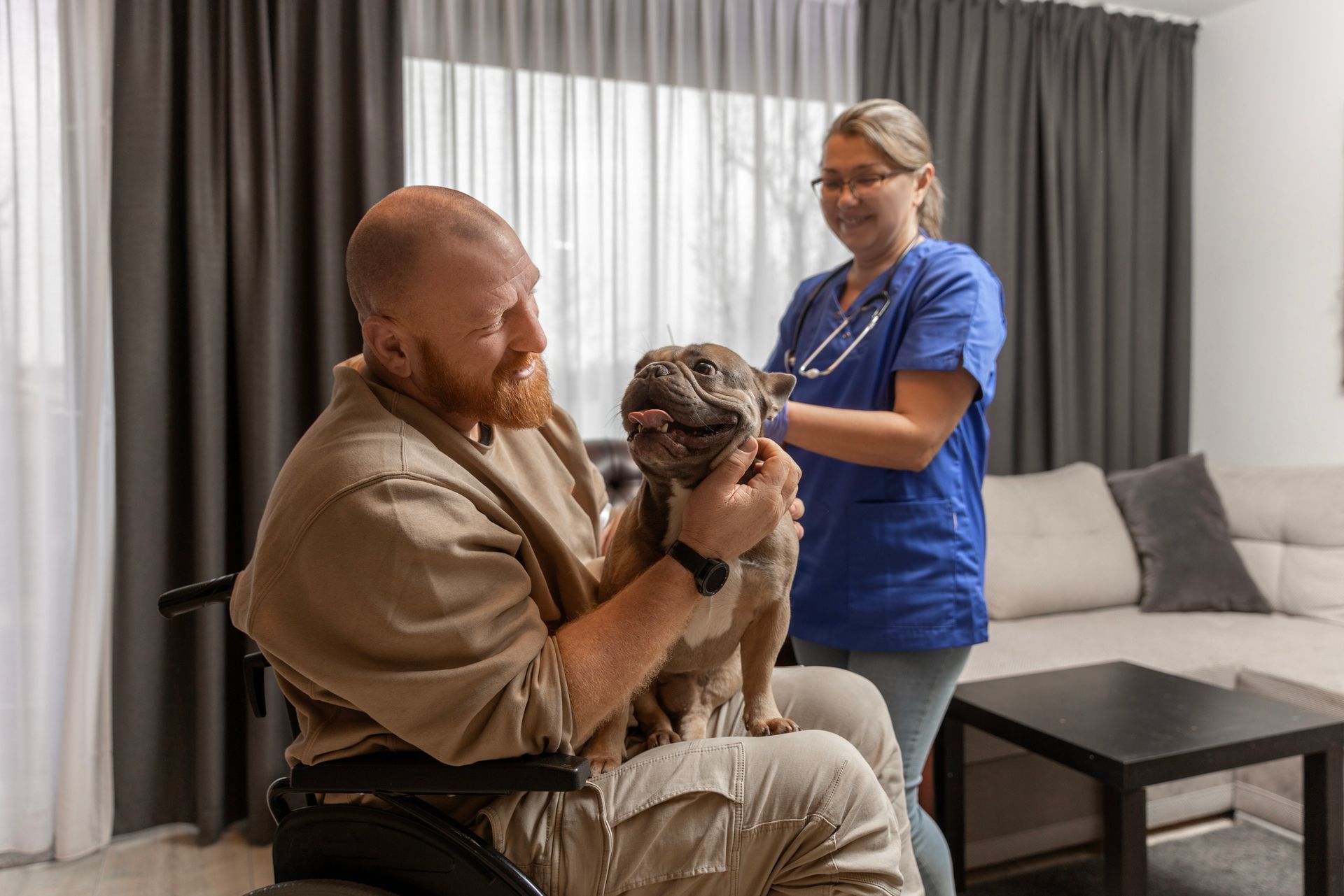 Man in wheelchair holds a dog as a vet checks it in a living room.