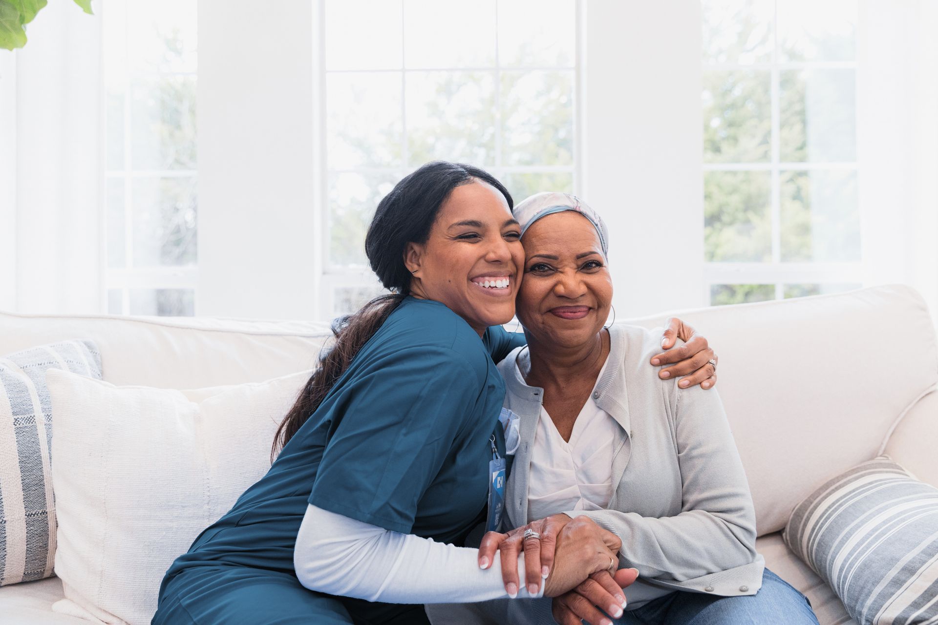 A smiling Black caregiver hugs an elderly Black woman on a white couch; both look happy.