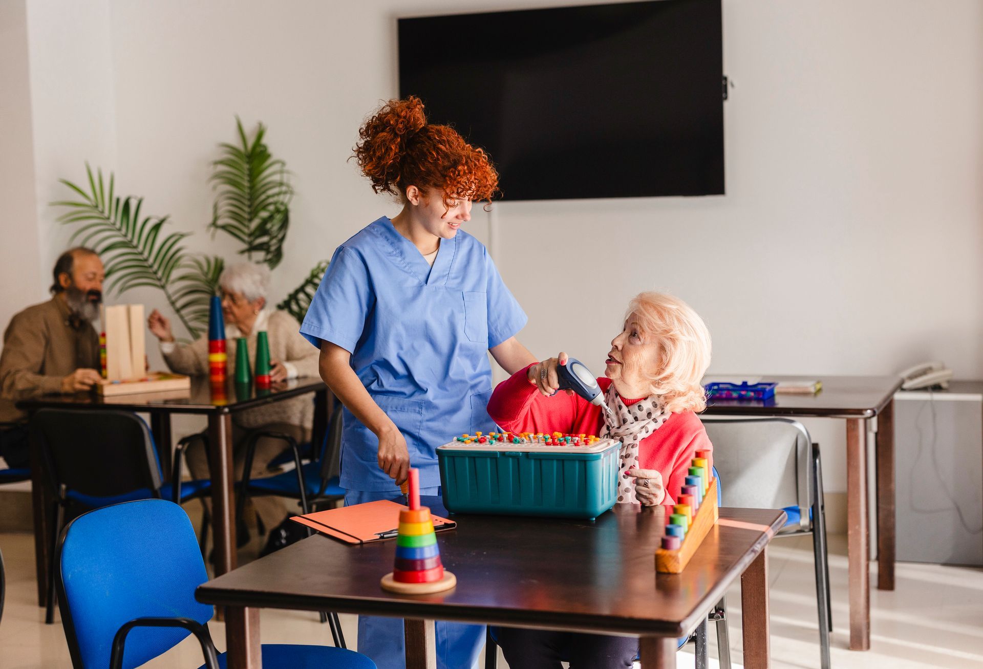 Caregiver in blue scrubs assisting an elderly woman with a cognitive game at a table; other seniors in background.