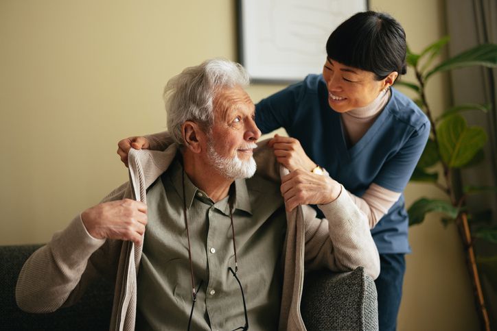 Woman in blue scrubs helps elderly man put on sweater.