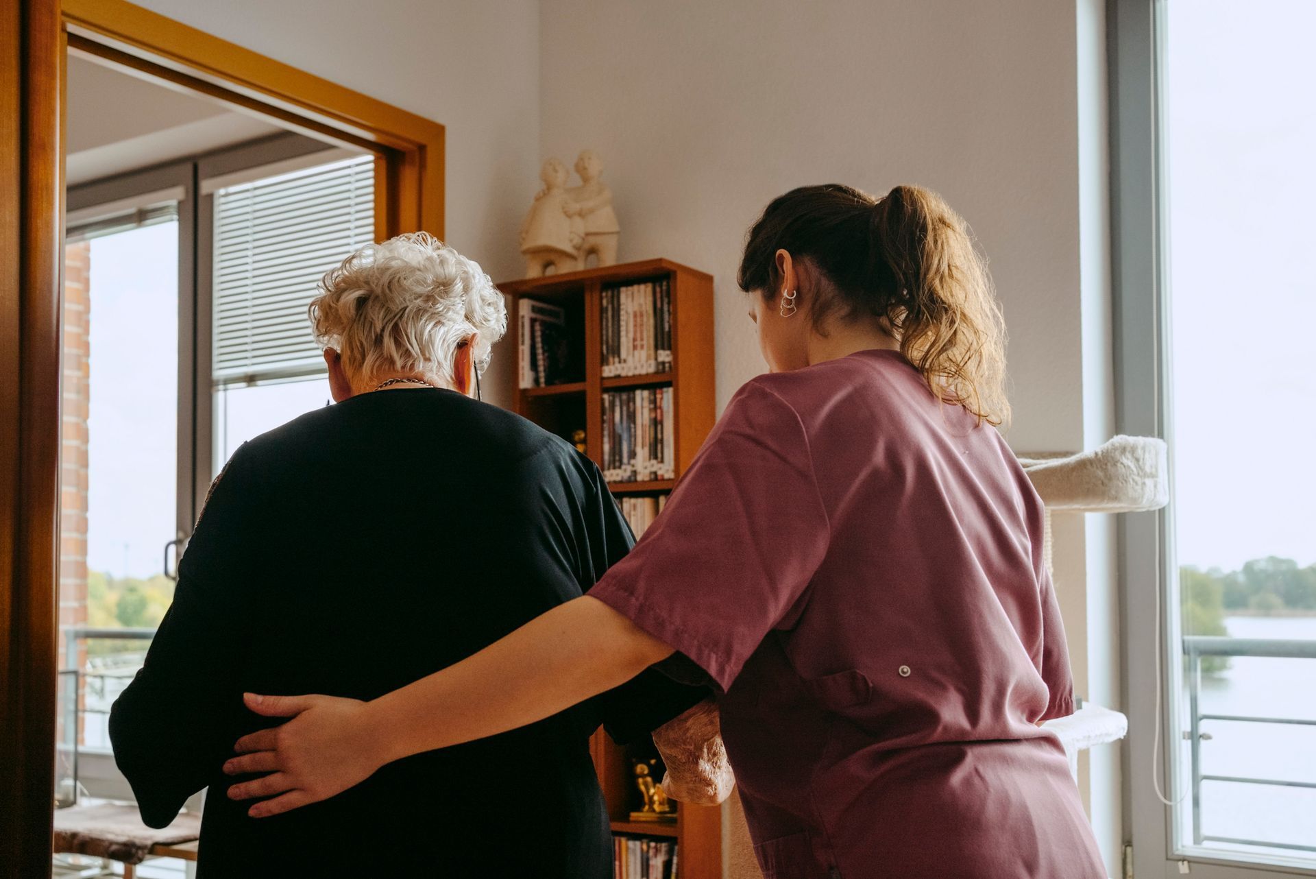 Caregiver assists an elderly person walking near a bookshelf and window, overlooking a body of water.