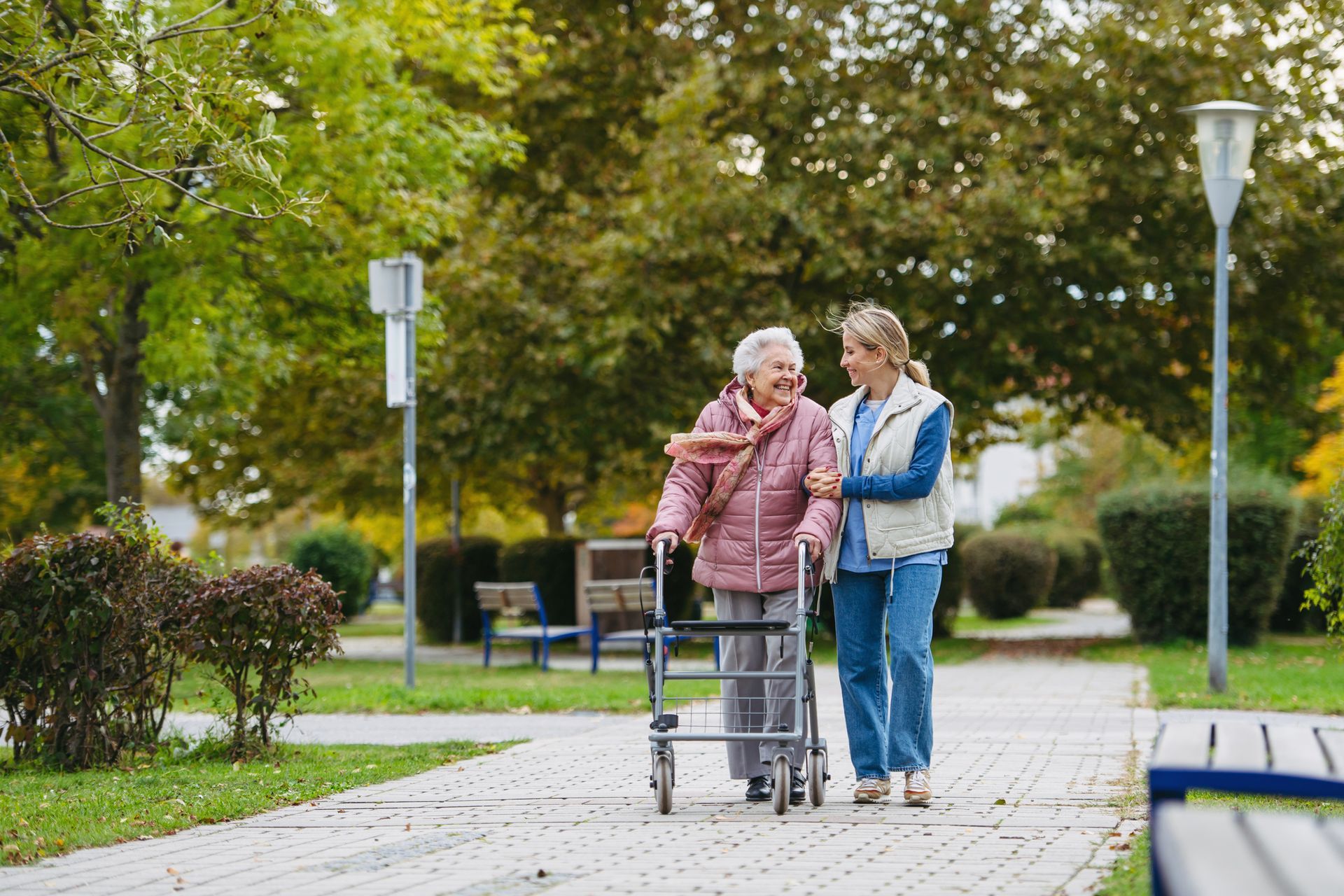 Elderly woman using a walker, assisted by a caregiver, walking outdoors on a path.