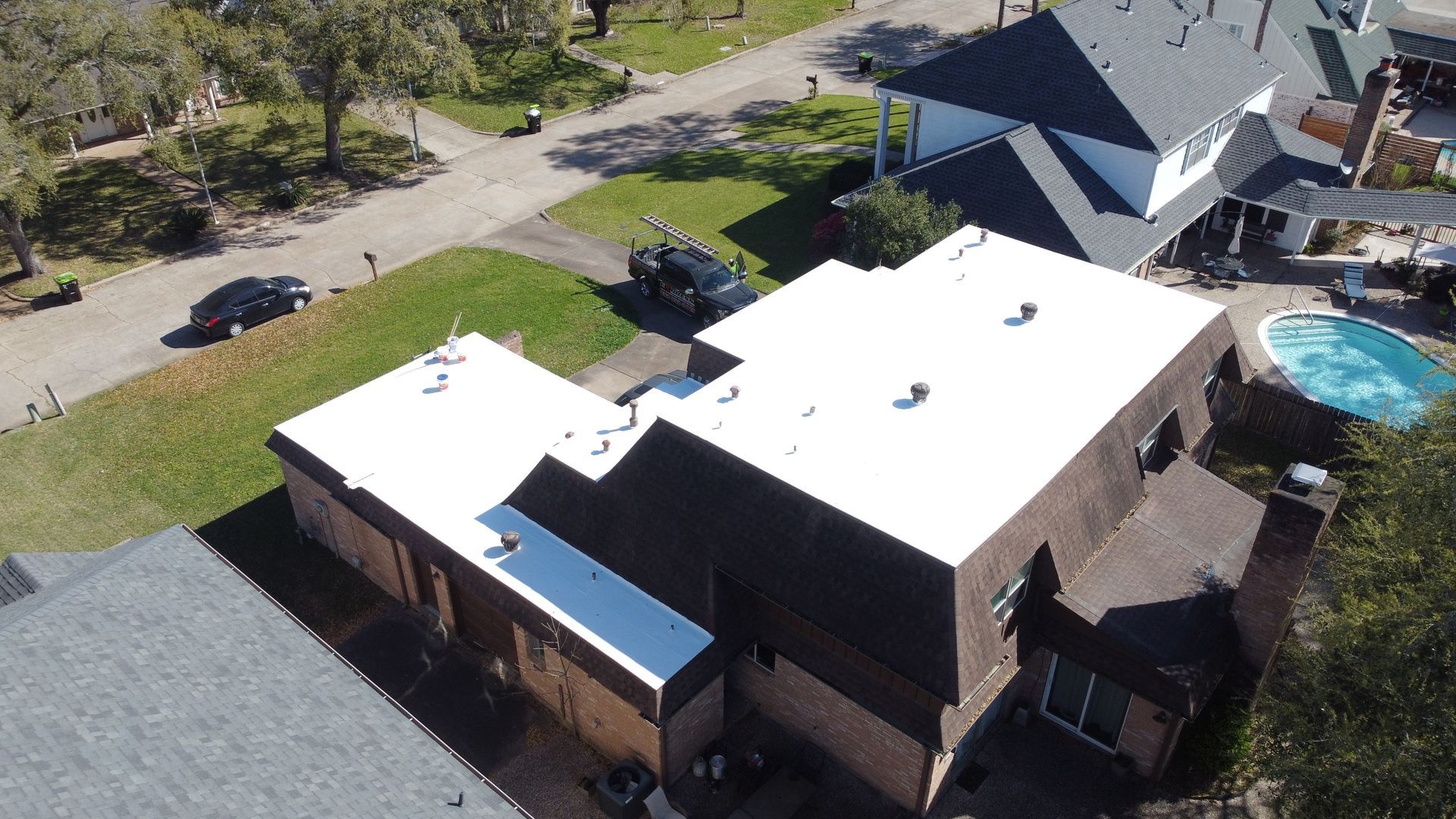 Aerial view of houses with white roofs and a swimming pool, cars on a road, trees and green lawn.