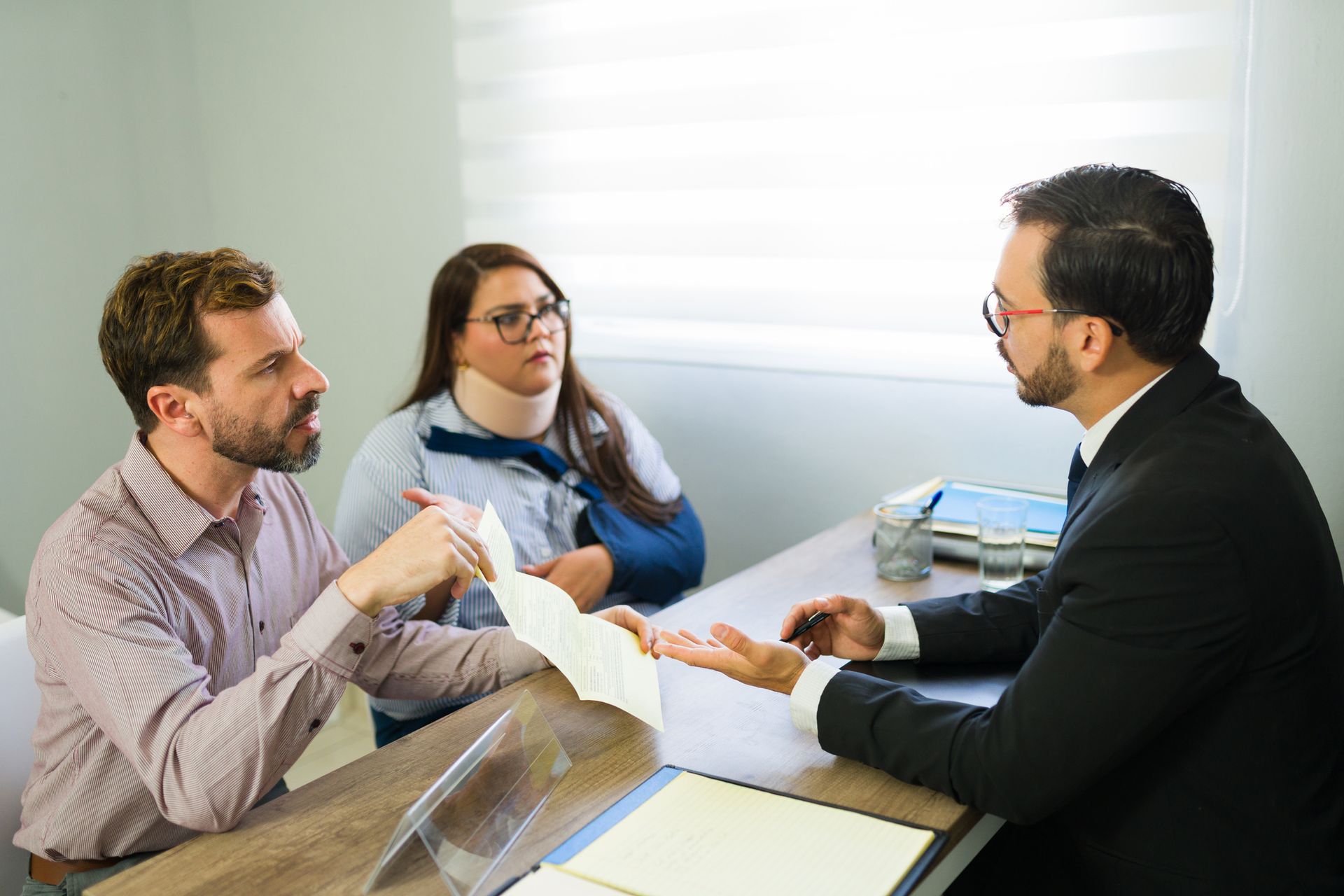 A couple and a lawyer discussing documents at a table. The woman has a neck brace.