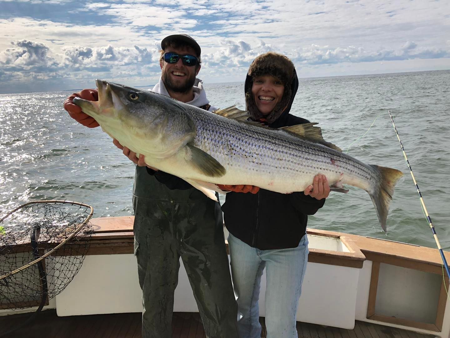 Two people on a boat hold a large striped fish over the water under a cloudy sky.