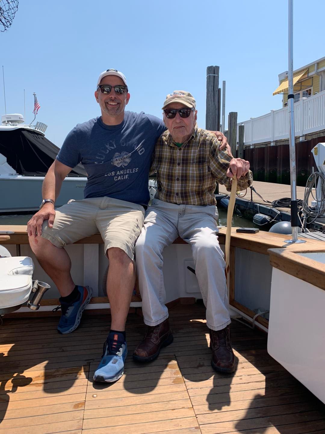 Two people sitting on a boat deck, smiling in the sun, with docks and boats in the background