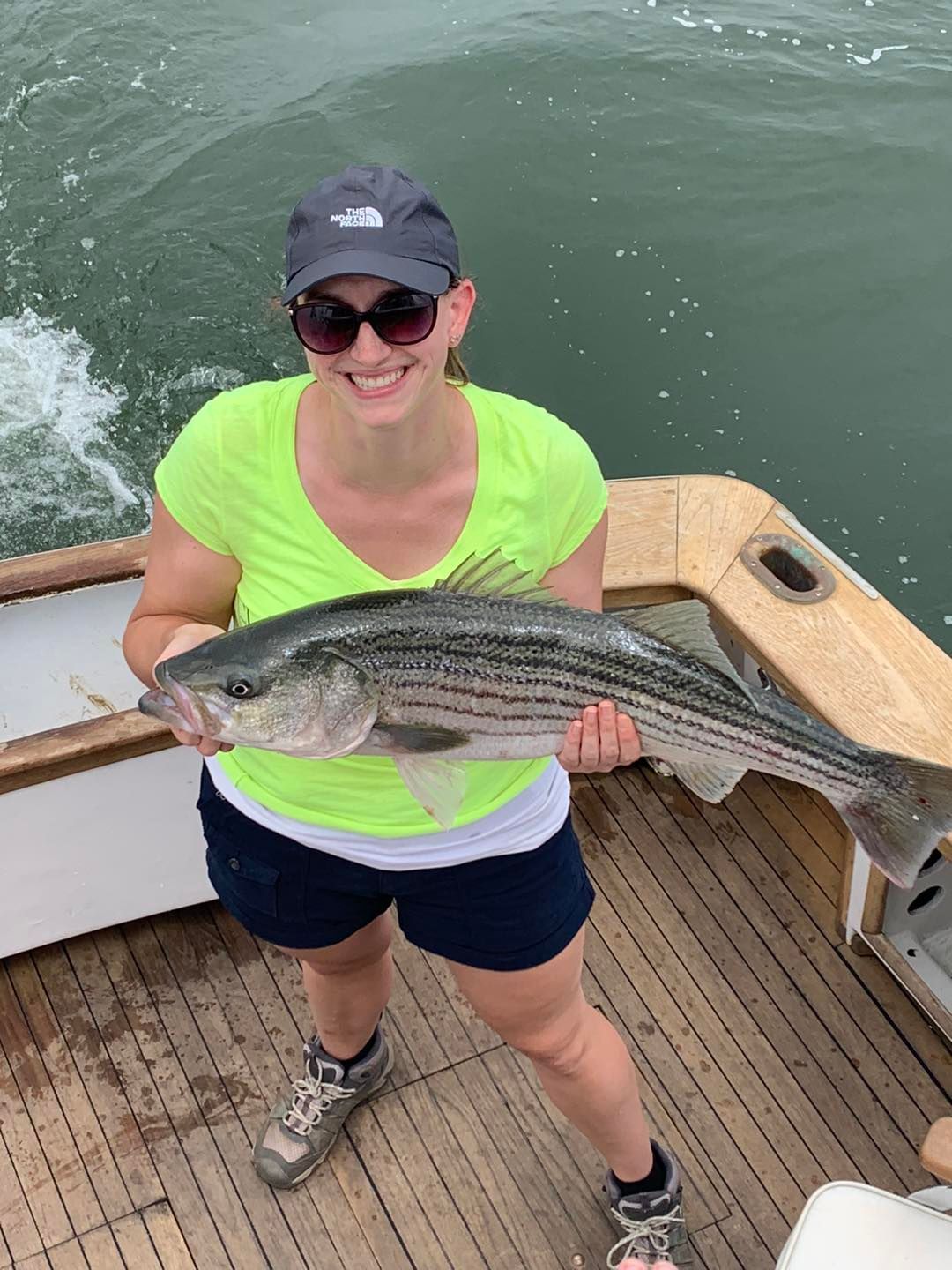 Person in a neon green shirt and cap smiles while holding a large striped fish on a boat deck.