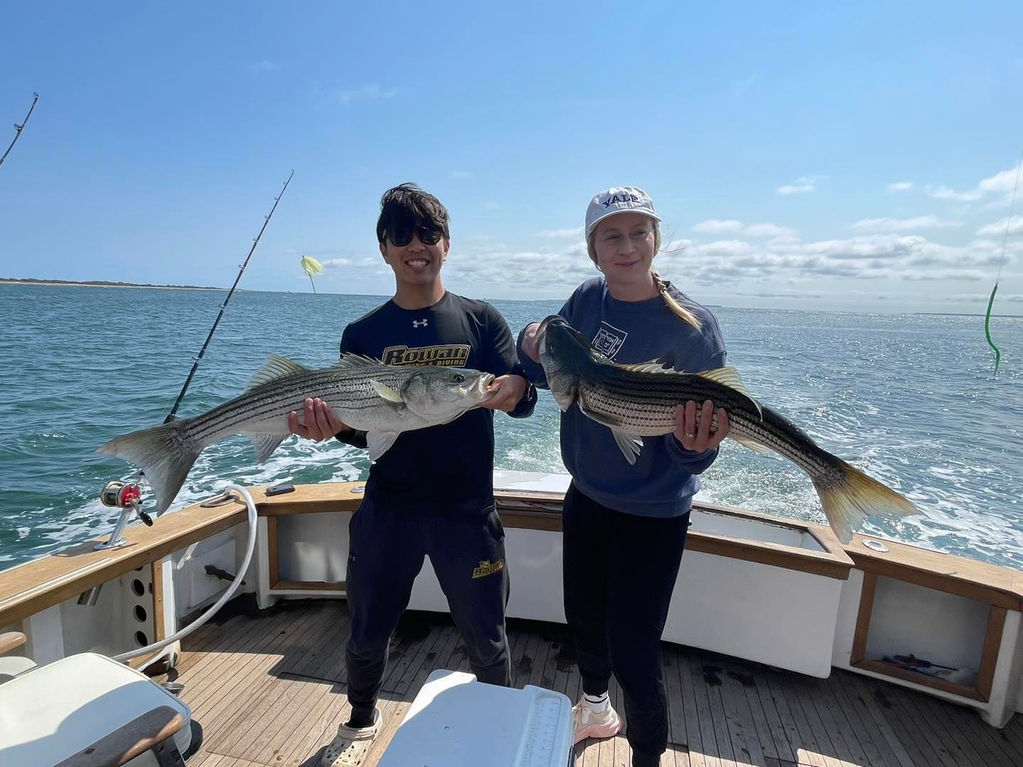 Two people holding large fish on a boat deck with fishing rods and calm blue water behind them