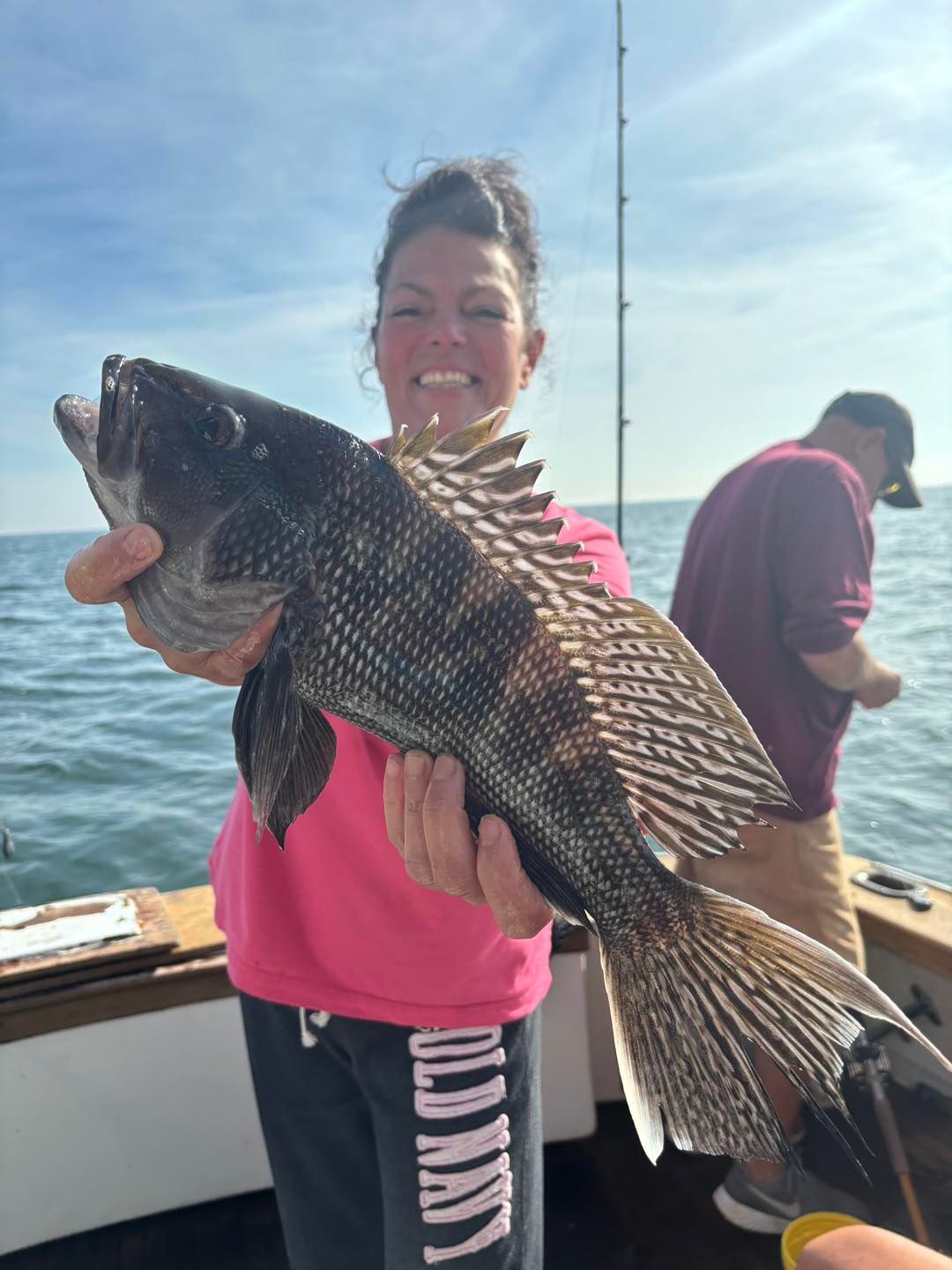 Smiling person holding a large striped fish on a boat, with another person in the background.