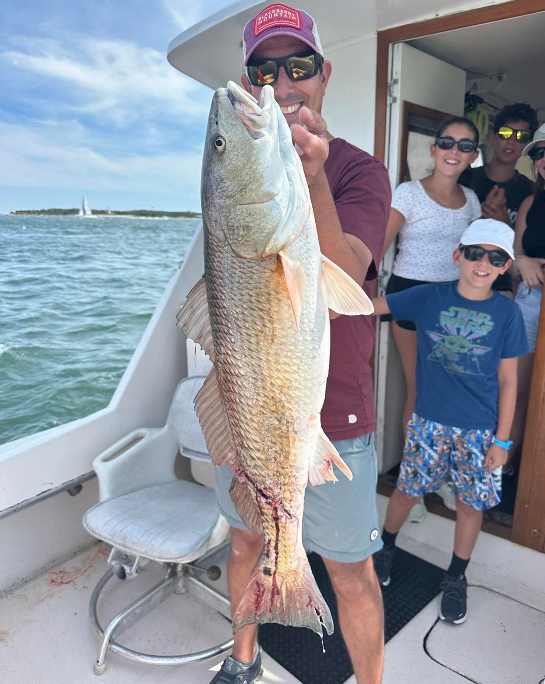 Man holding a large silver fish on a boat, with two smiling people standing behind him on the deck.