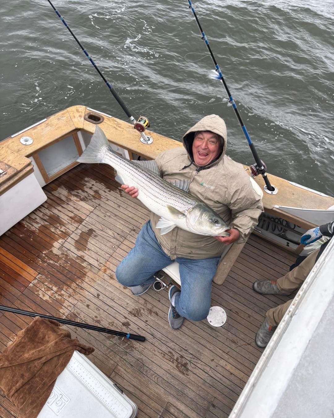 Person kneeling on a boat deck, holding a large striped fish and smiling on the water.