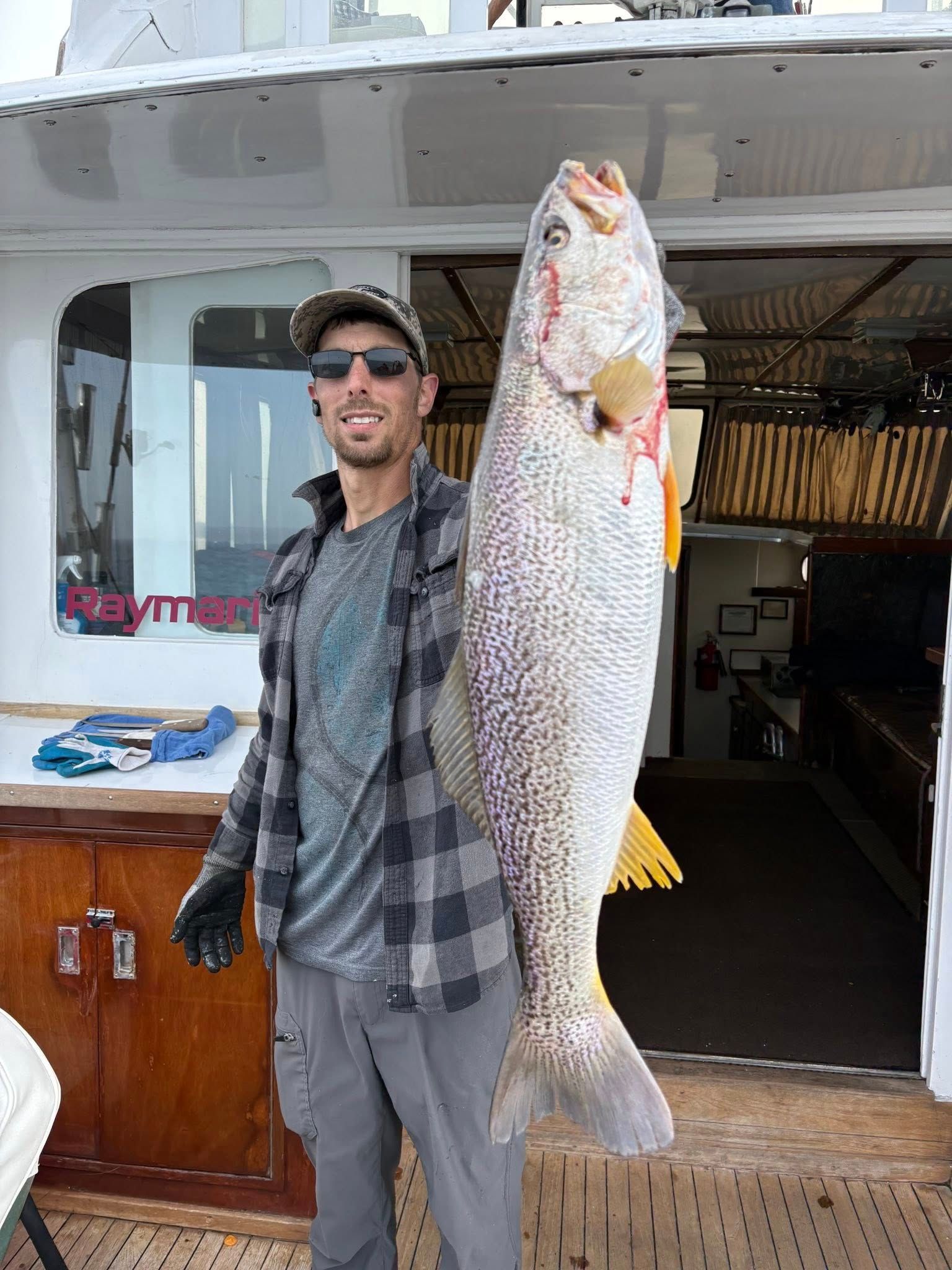 Man holding a large silver fish on a boat deck, standing by the cabin doorway.