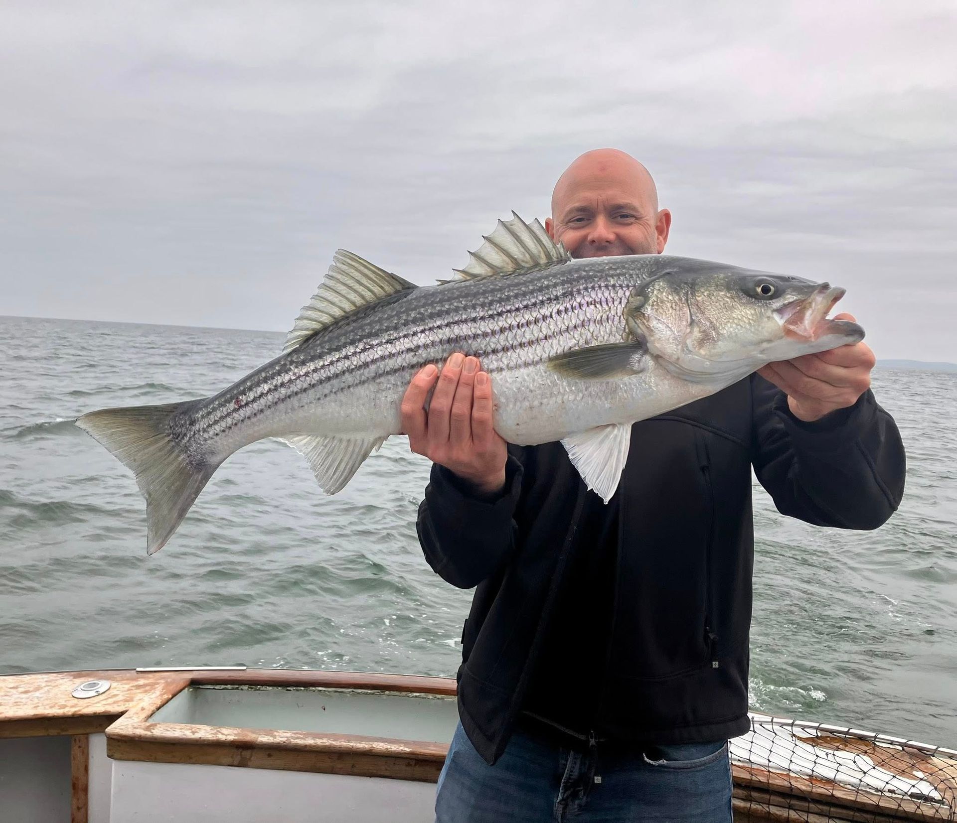 Man holding a large striped bass on a boat, with choppy water and overcast sky behind him