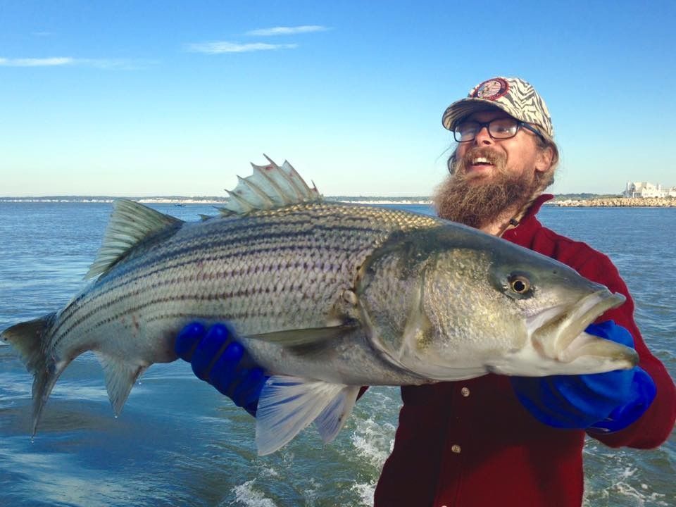 A person with a beard and patterned cap holds a large, striped fish against a background of water and a bright blue sky.