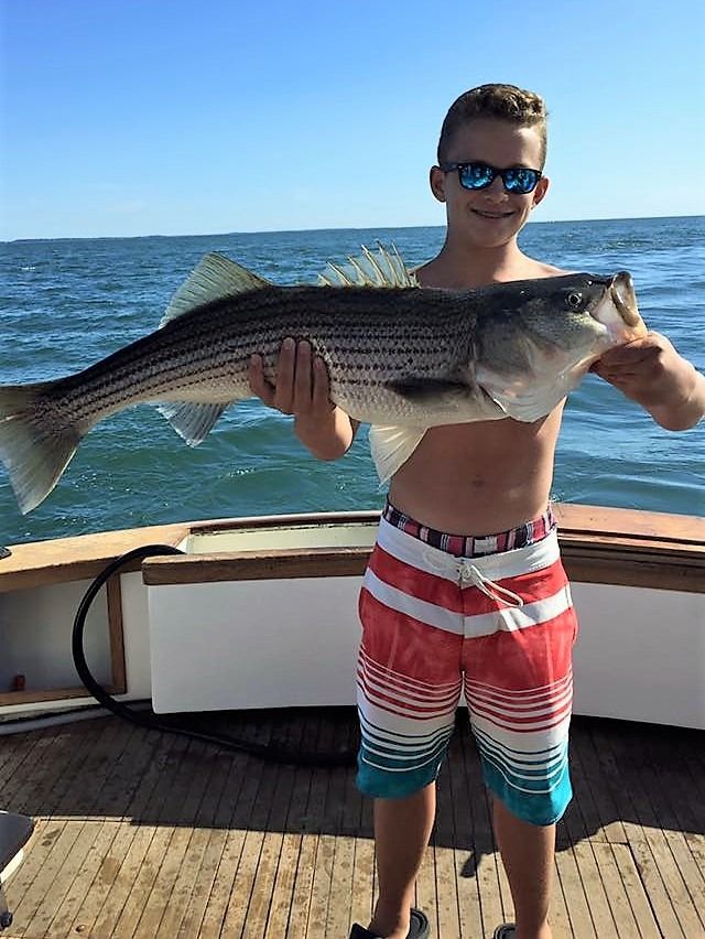A person wearing sunglasses and patterned swim trunks stands on a boat deck holding a large, striped fish.