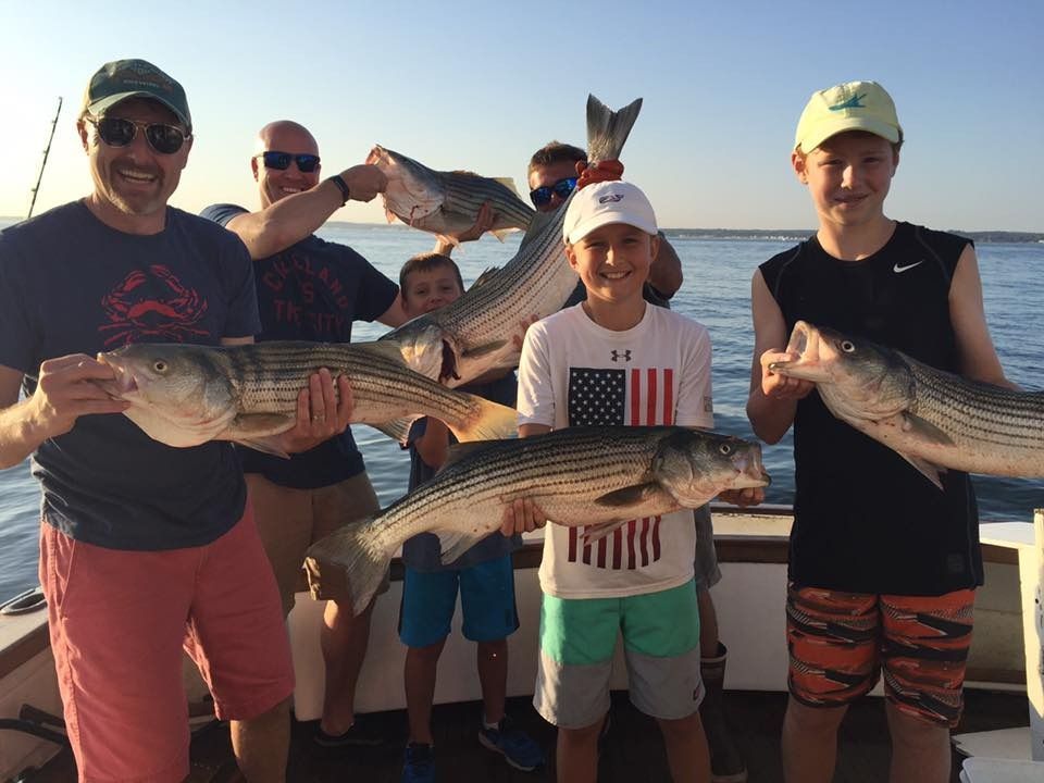 Group of people on a boat holding large striped fish caught during a fishing trip on a sunny day.