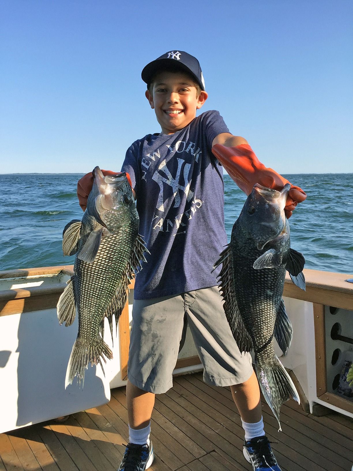 Boy on a boat deck holding two large fish, smiling by the water.
