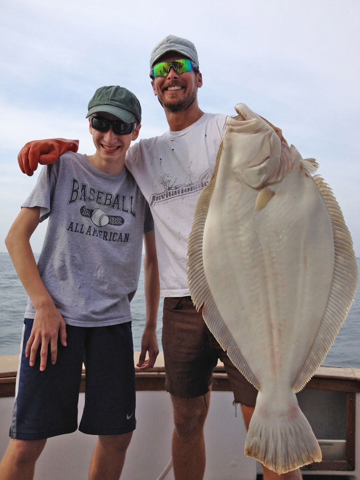 Two people on a beach holding a large fish, both smiling and wearing hats and sunglasses.