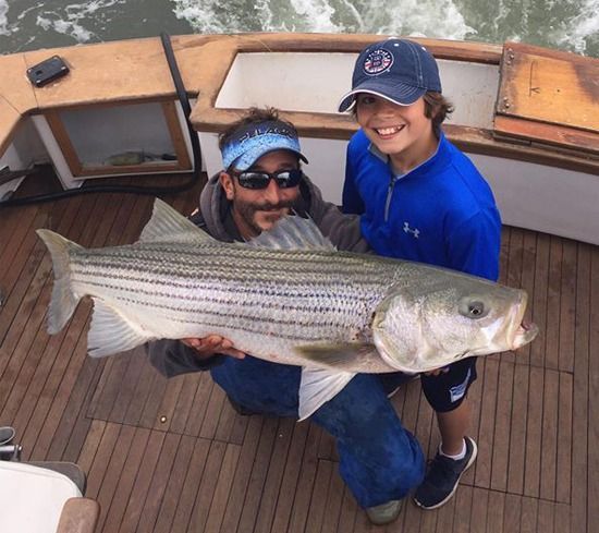 An adult and child holding a large striped bass on the wooden deck of a boat.