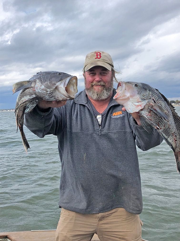 Man on a boat holding two large fish, with cloudy sky and water in the background.