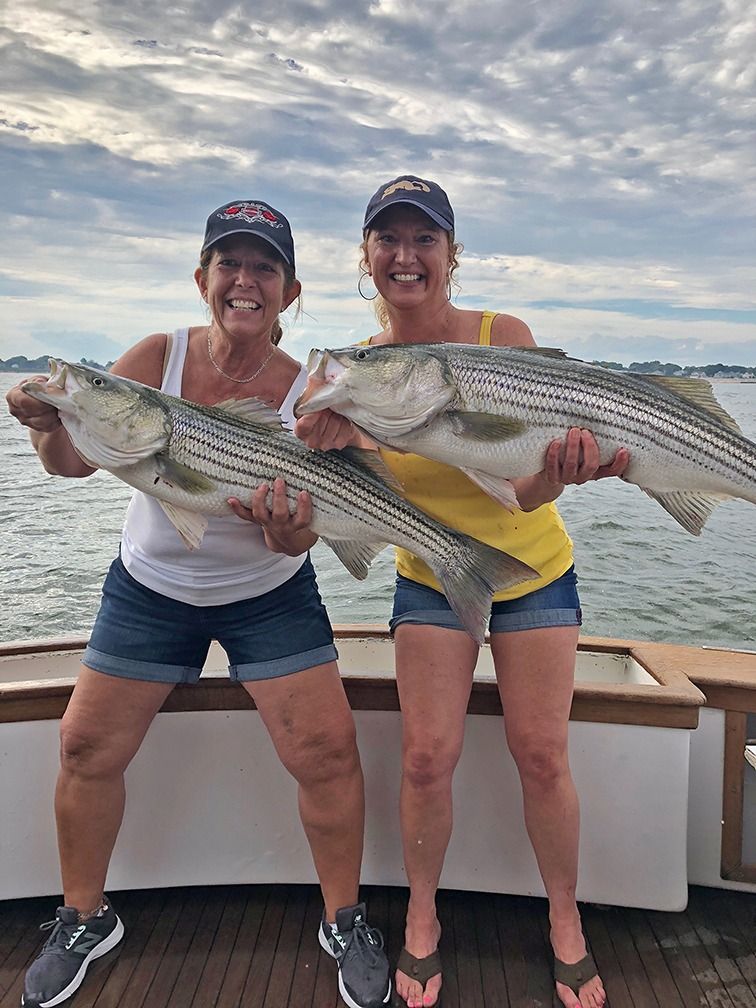 Two people on a boat smiling and holding large striped fish at sunset.