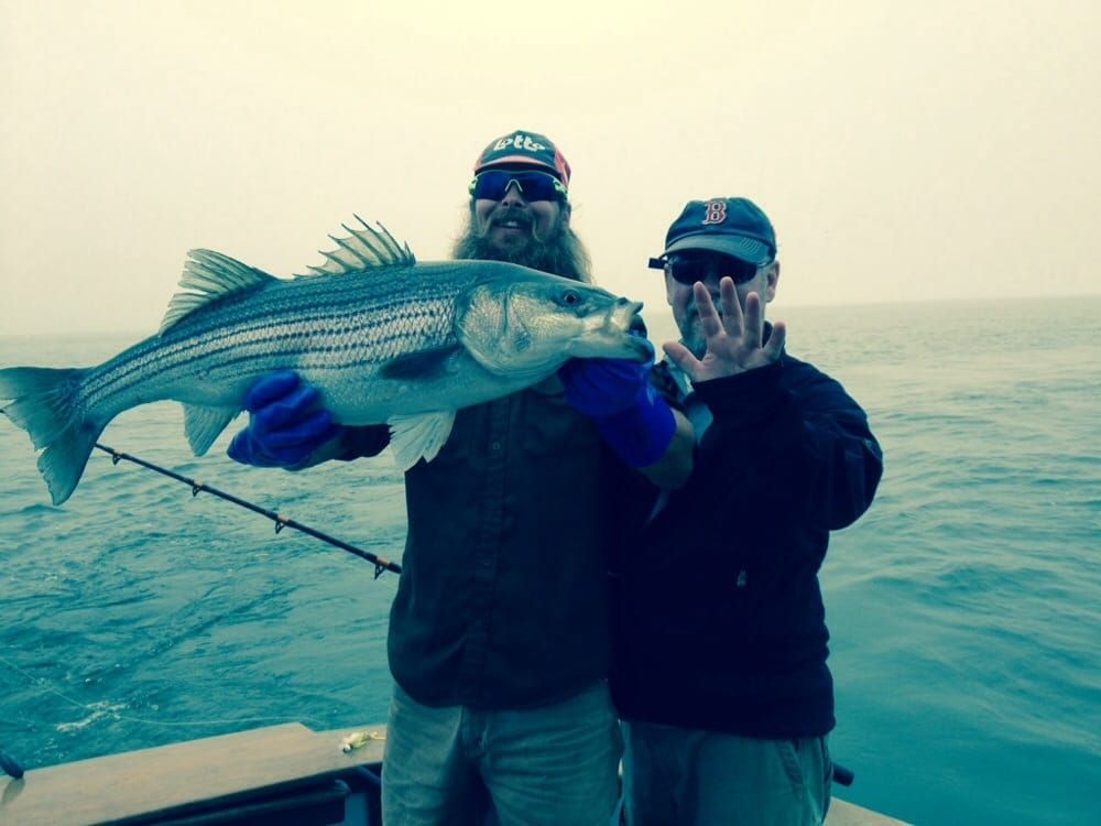 Two people on a boat hold up a large striped fish against the ocean backdrop