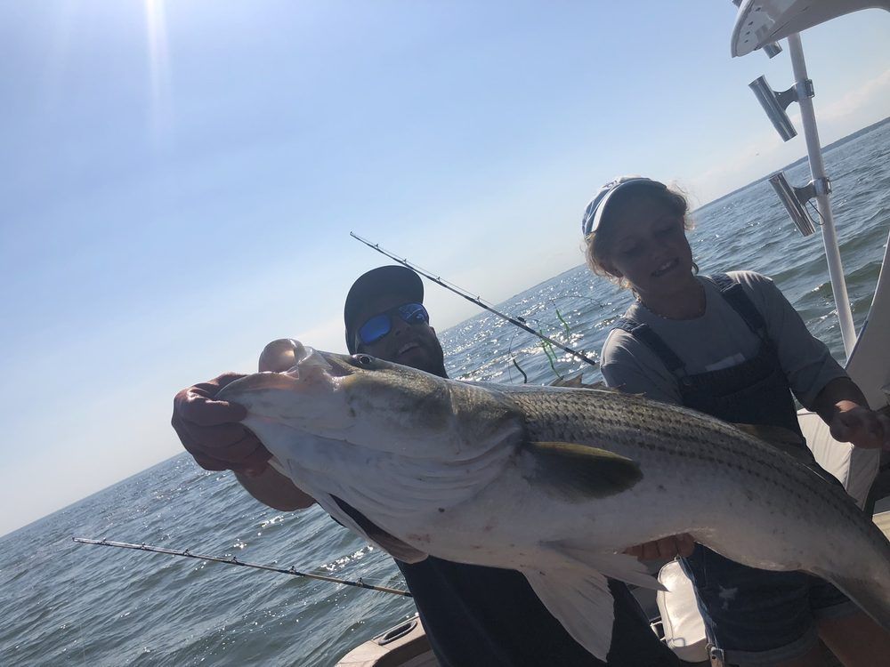 Two people on a boat hold a large striped fish over open water under a cloudy sky