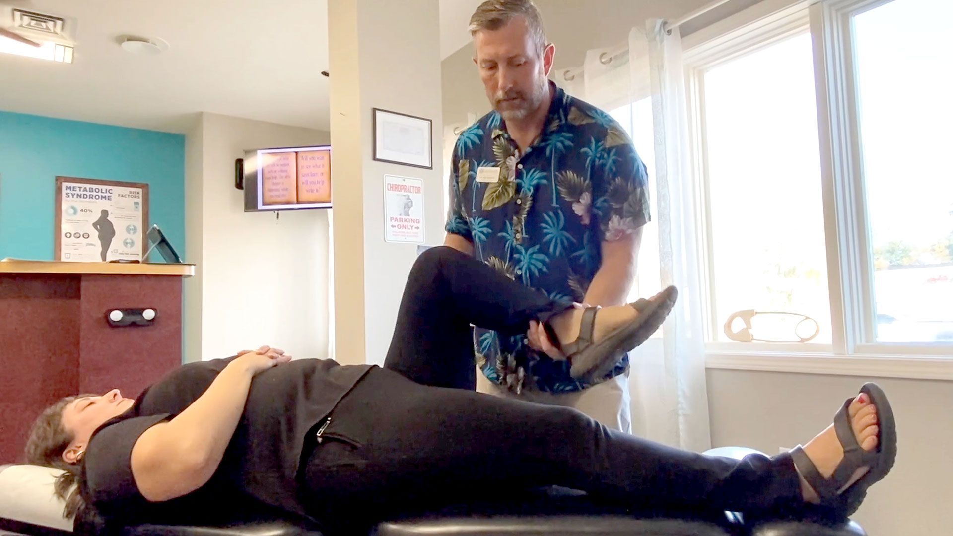 Person lying on a table while a person in a floral shirt examines their leg in a clinic.