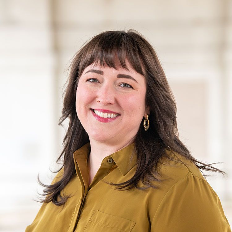 Woman with dark hair and bangs smiles, wearing a gold shirt, against a blurred, light background.