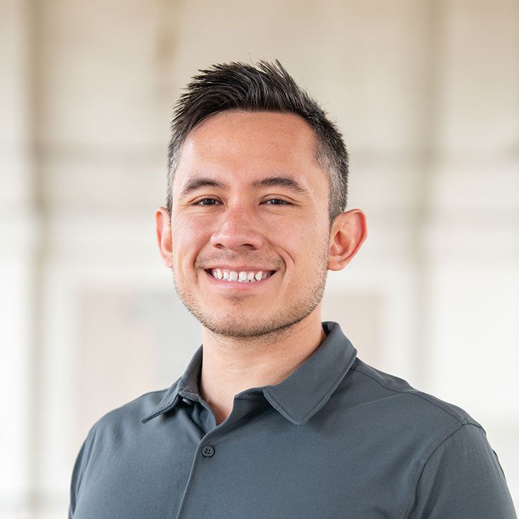Man in gray shirt smiling, headshot. Blurred, bright background.