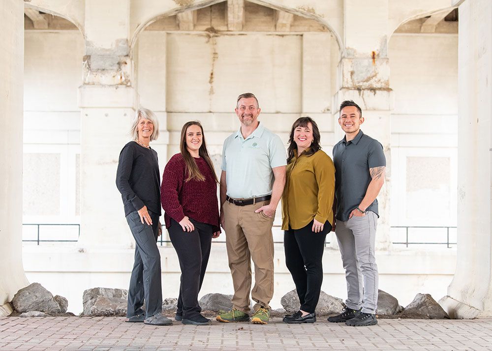 Five people stand under a white concrete structure. They are smiling, and looking at the camera.