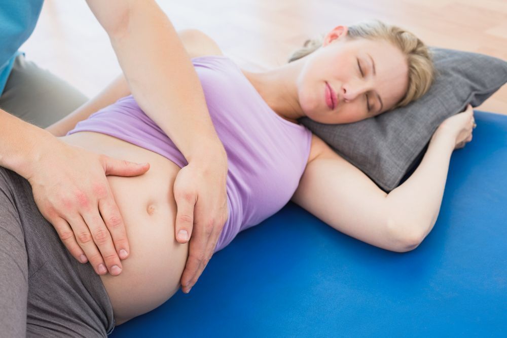 Pregnant woman receiving a massage on a blue exercise ball; person's hands on her belly.