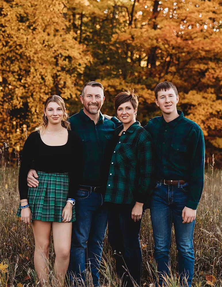 Family of four poses outdoors in fall, wearing green and denim with golden foliage backdrop.
