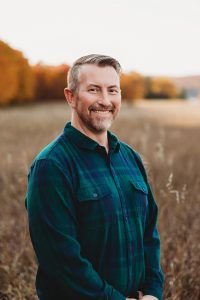 Man in green plaid shirt smiling outdoors in field with fall foliage.