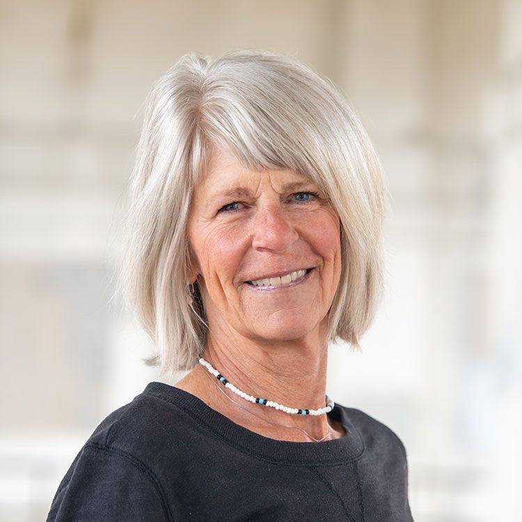 Woman with short, gray hair smiles, wearing a black shirt and a beaded necklace. Blurred, light background.
