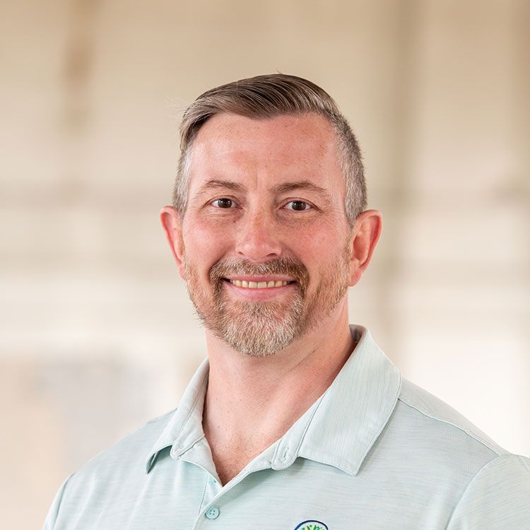 Man with a short beard smiles, wearing a light blue collared shirt. Soft, neutral background.