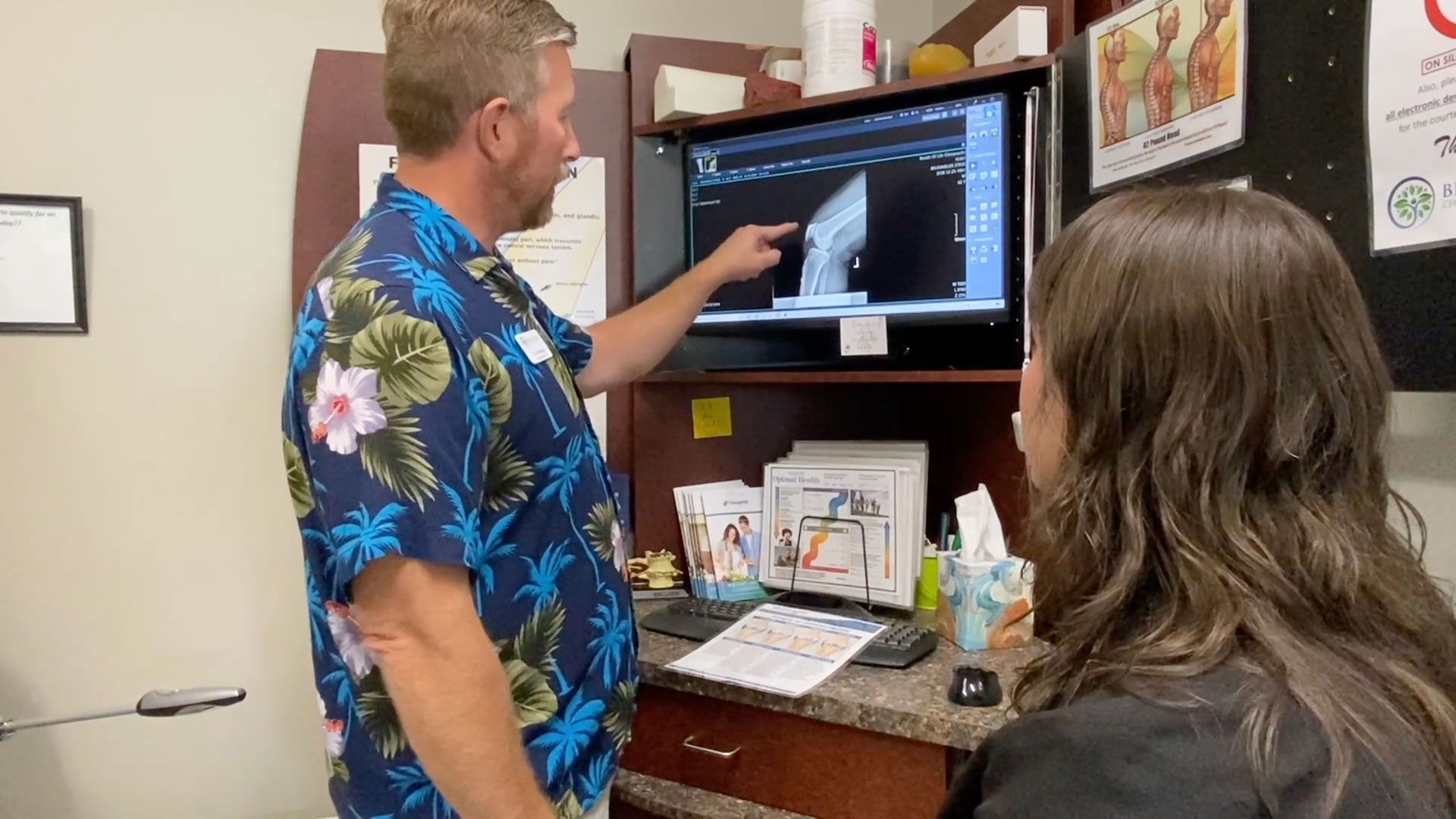 Man in floral shirt points to ultrasound image on a monitor, showing a woman the results in an office.