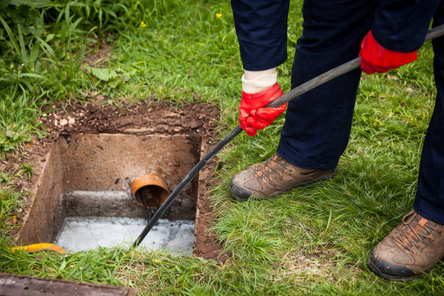 A worker in red gloves uses a cable to clear a blockage in an open residential drainage inspection chamber.