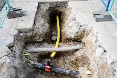 A trench cut into a concrete sidewalk reveals a yellow gas pipe connected to a black main line and a grey conduit.