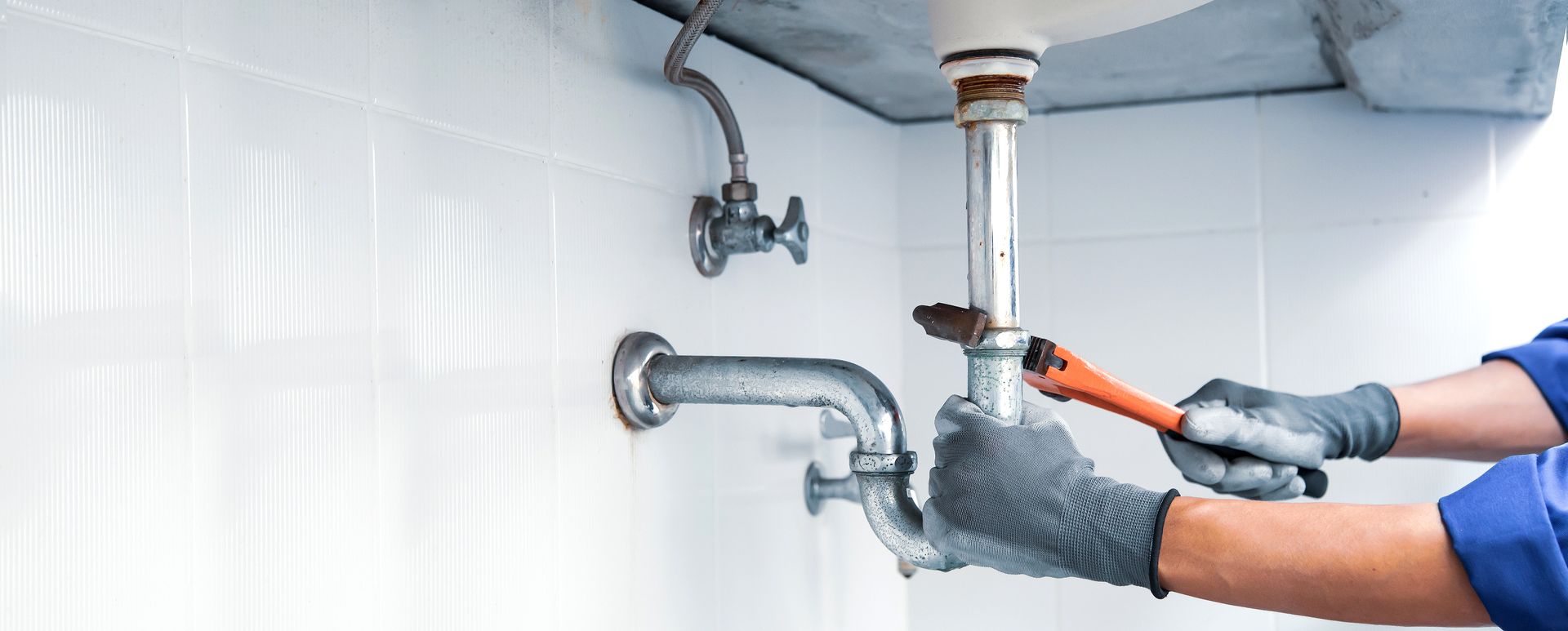 A plumber wearing work gloves uses a pipe wrench to tighten a metal pipe beneath a bathroom sink.