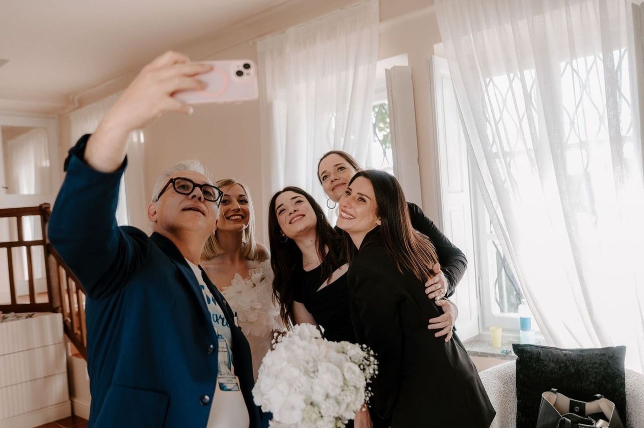 Gruppo di donne che si scattano un selfie in un interno: la sposa con il bouquet, altre sorridenti, tende leggere e finestra.