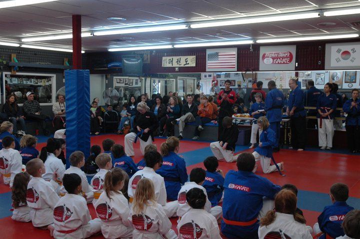 A group of kids in karate uniforms are sitting on the floor in a gym