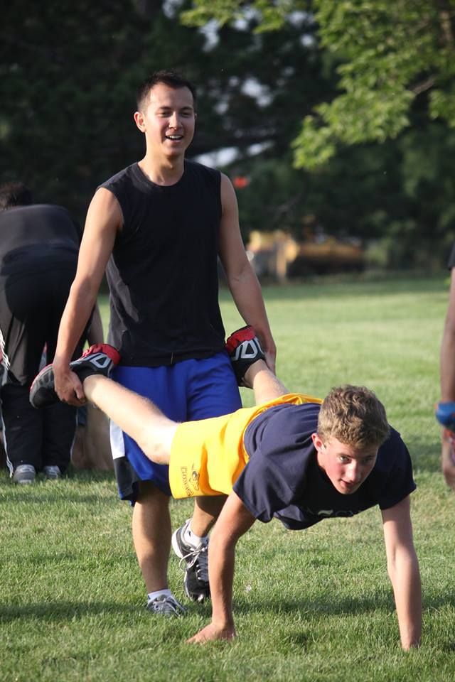 A man is helping another man do push ups on the grass