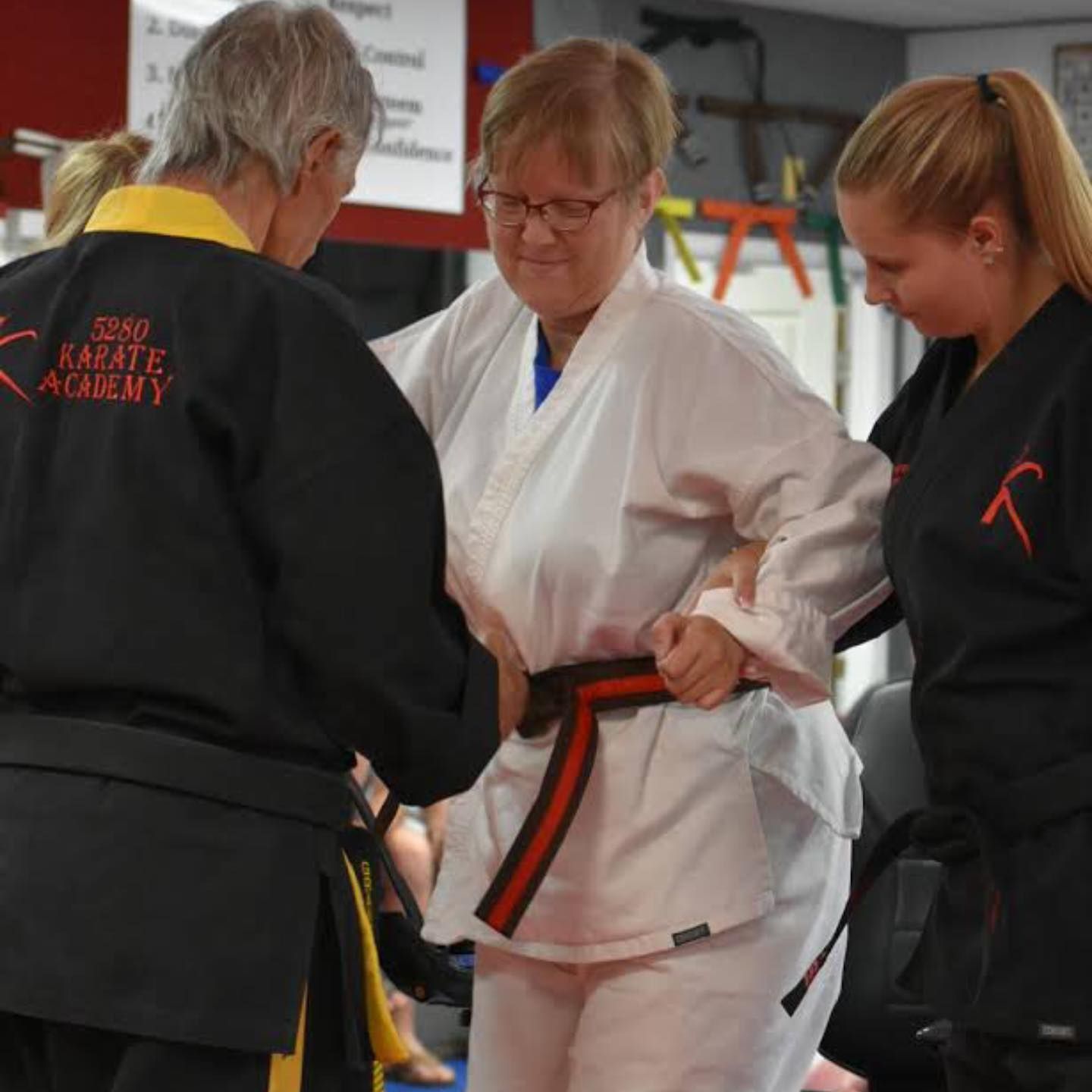 A man in a black karate academy uniform helps a woman with her belt