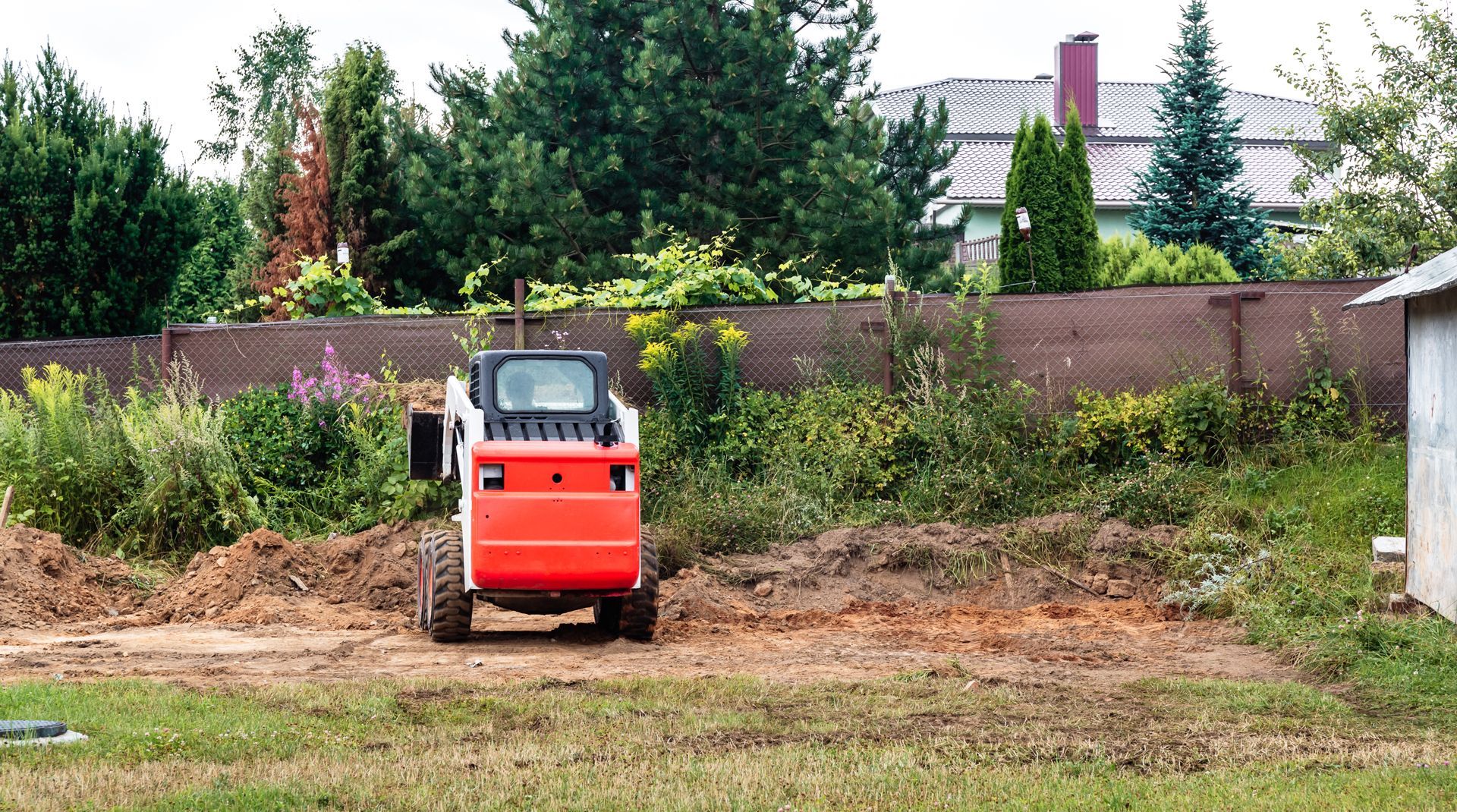 Orange Bobcat skid-steer loader on dirt clearing, near greenery and a fence.