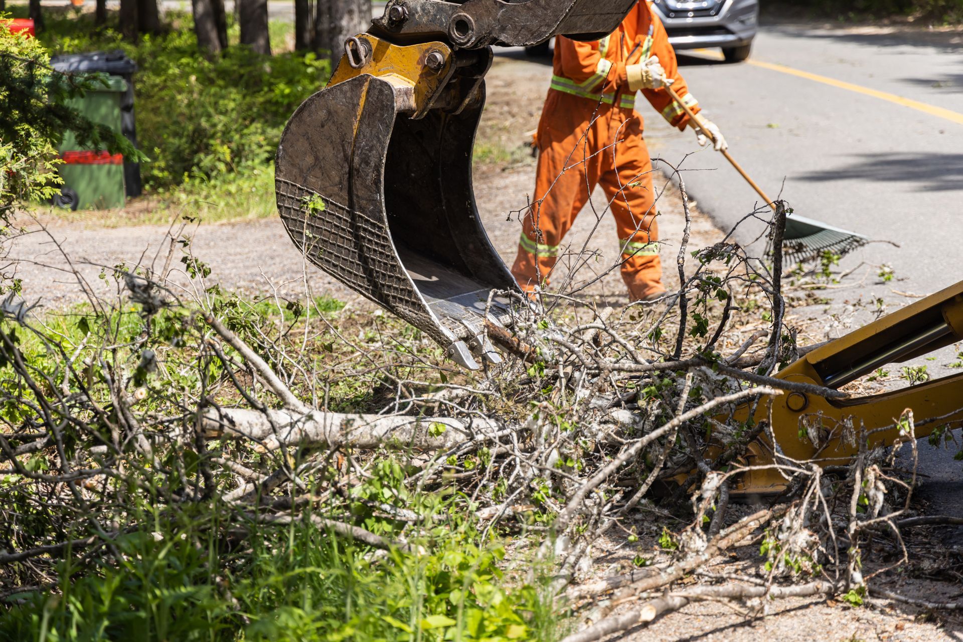 A person in orange work clothes using an excavator bucket to clear brush along a road.