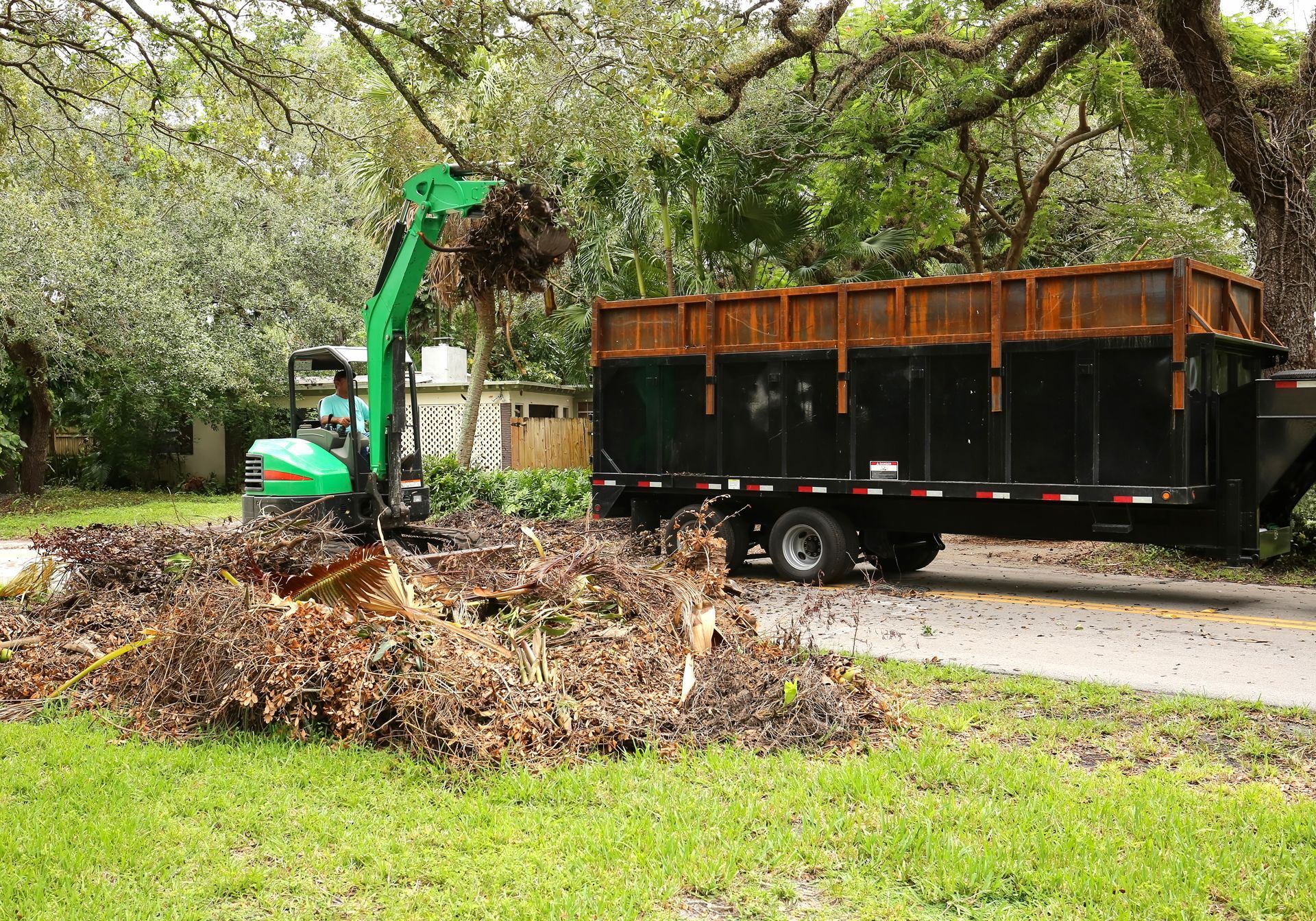 Green wood chipper loading debris into a trailer on a tree-lined street.
